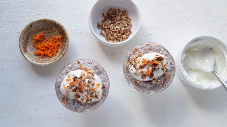 Carrot cake chia pudding in bowls next to bowl of shredded carrots, chopped pecans, and yogurt