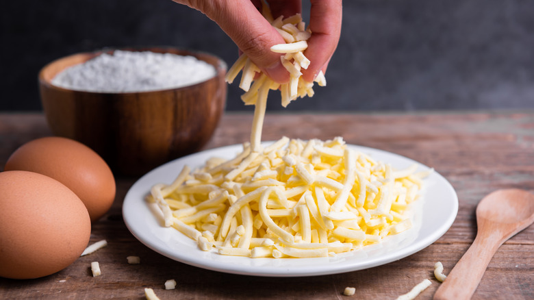 A person holds a handful of grated mozzarella cheese above a plate